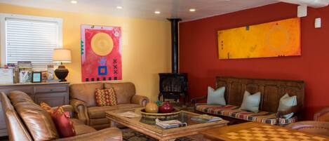 A living room with a sofa, armchairs, a coffee table, and a chess table. There is a fireplace, a window with blinds, and artwork on the walls.