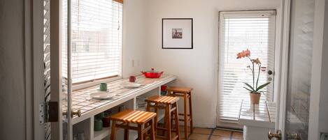 A kitchen with a tiled backsplash, a window with blinds, a red pot on the counter, and a potted orchid by the door.