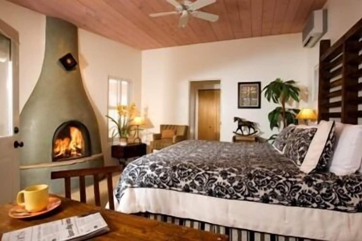 A bedroom with a fireplace, a bed with patterned bedding, a wooden table with a cup and a book, and a ceiling fan.