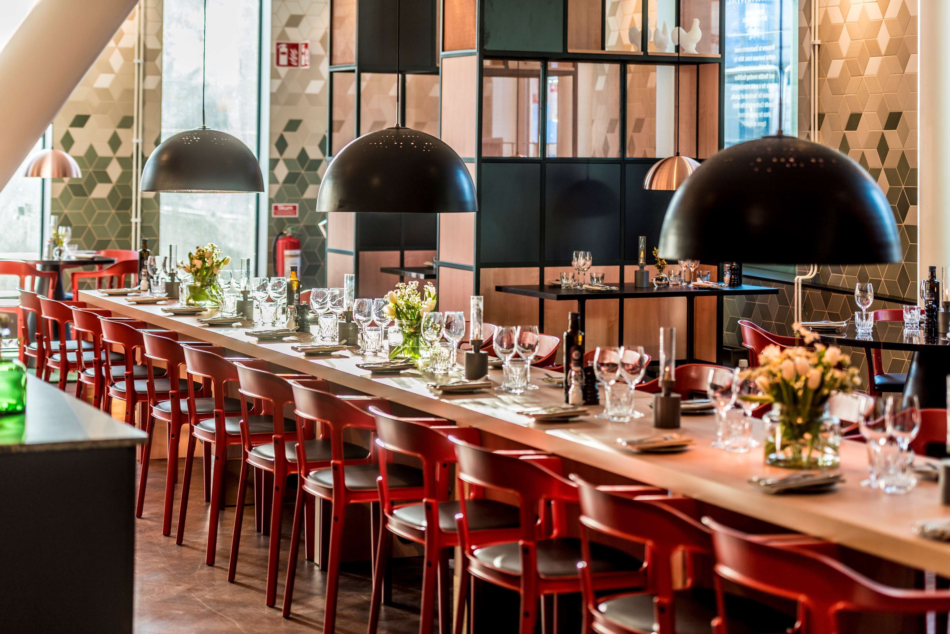 A restaurant with long tables, red chairs, black pendant lights, and a geometric wall pattern.