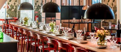 A restaurant with long tables, red chairs, black pendant lights, and a geometric wall pattern.