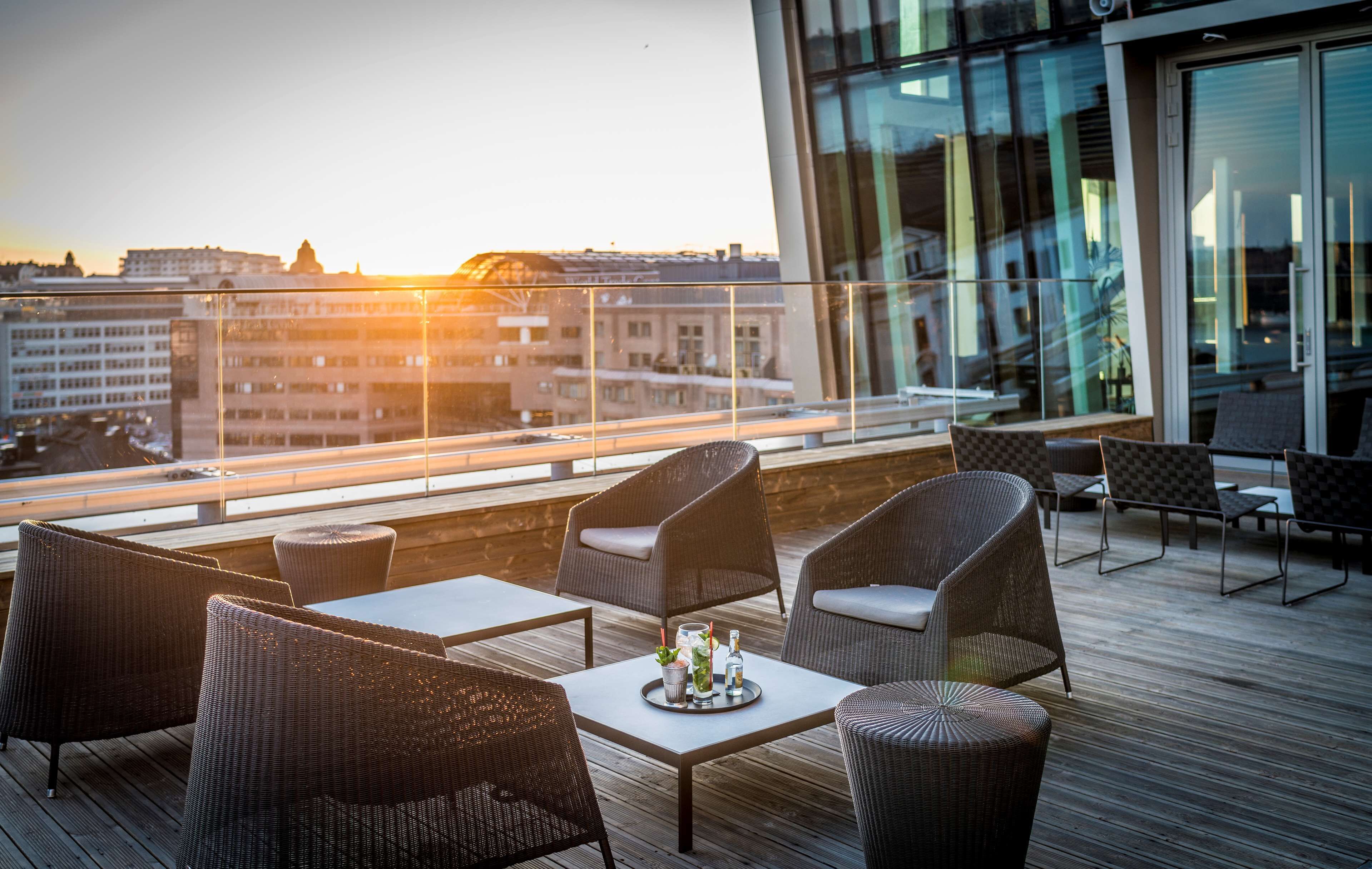 A rooftop terrace with wicker furniture, a table with a tray of drinks, and a city view at sunset.