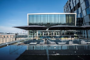 Área de piscina no terraço, com espreguiçadeiras e guarda-sóis, um edifício de vidro ao fundo e céu claro.