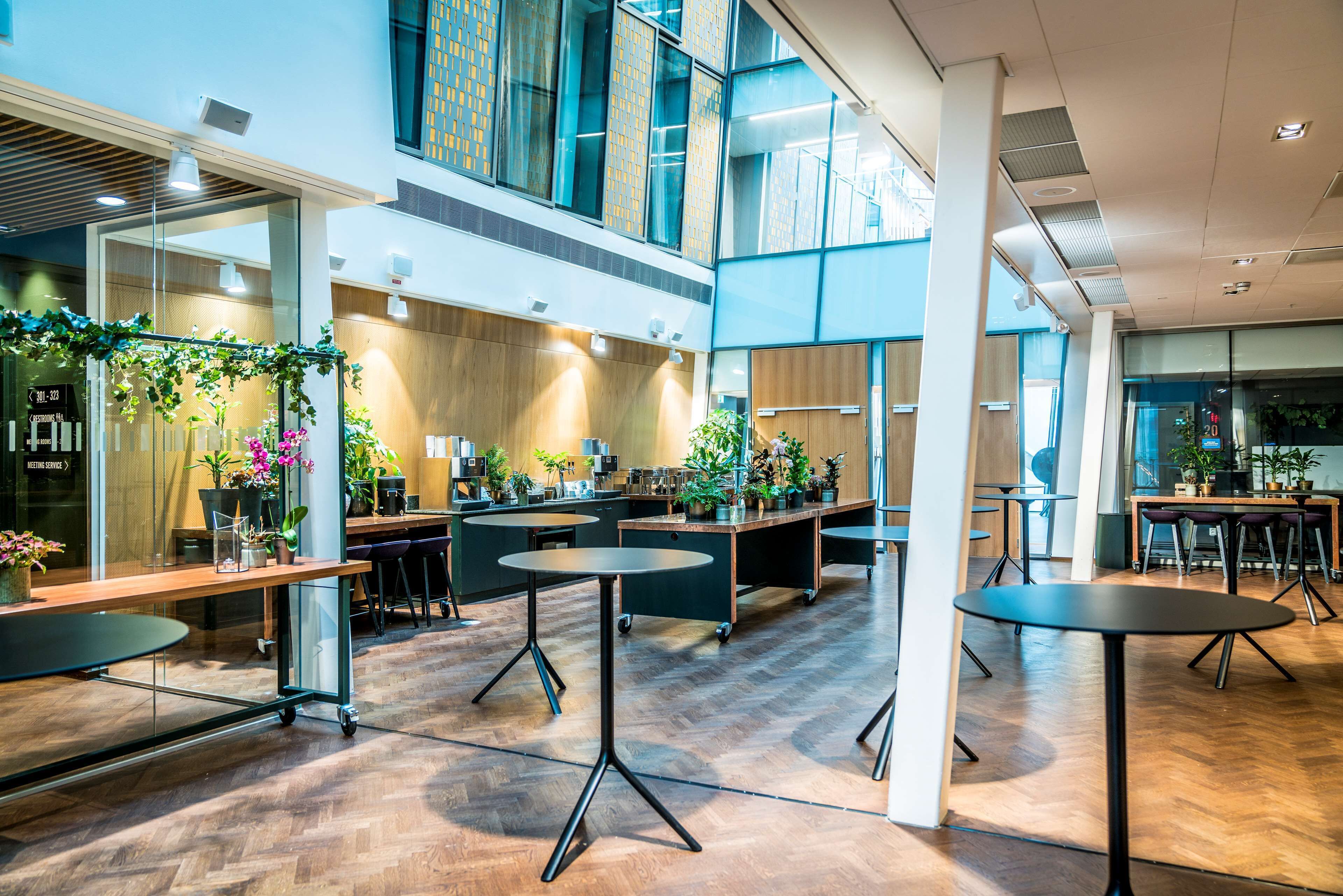 Een ruime, overdekte ruimte met meerdere tafels en stoelen, een keukenhoek en grote ramen voor natuurlijk licht.