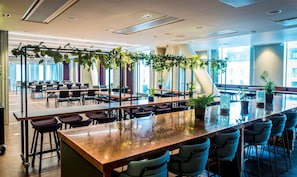 A spacious indoor area with multiple tables and chairs, large windows, and indoor plants.
