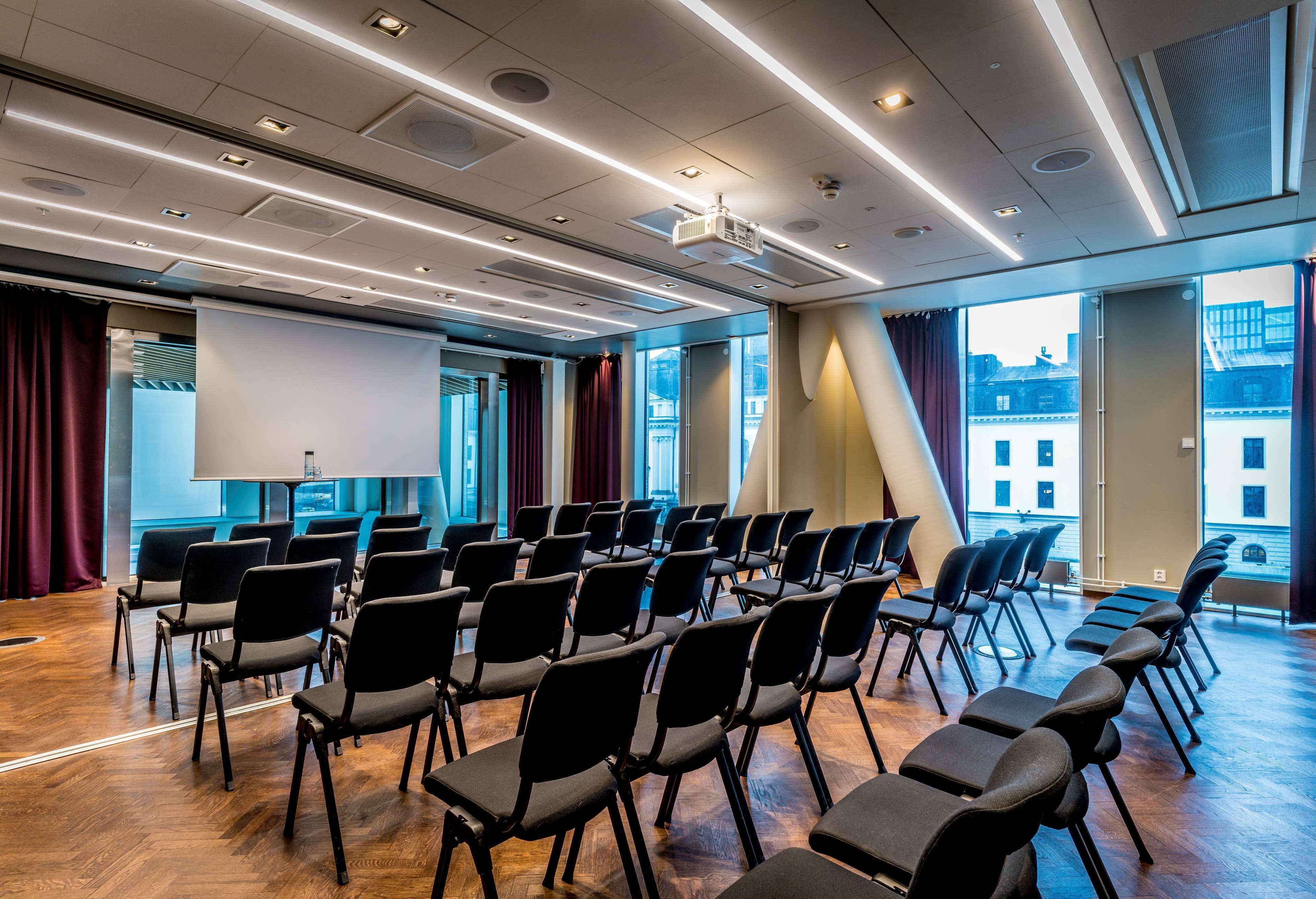 Un salón de conferencias con filas de sillas, una pantalla de proyector y ventanas amplias con vistas a la ciudad.
