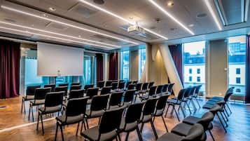 Un salón de conferencias con filas de sillas, una pantalla de proyector y ventanas amplias con vistas a la ciudad.