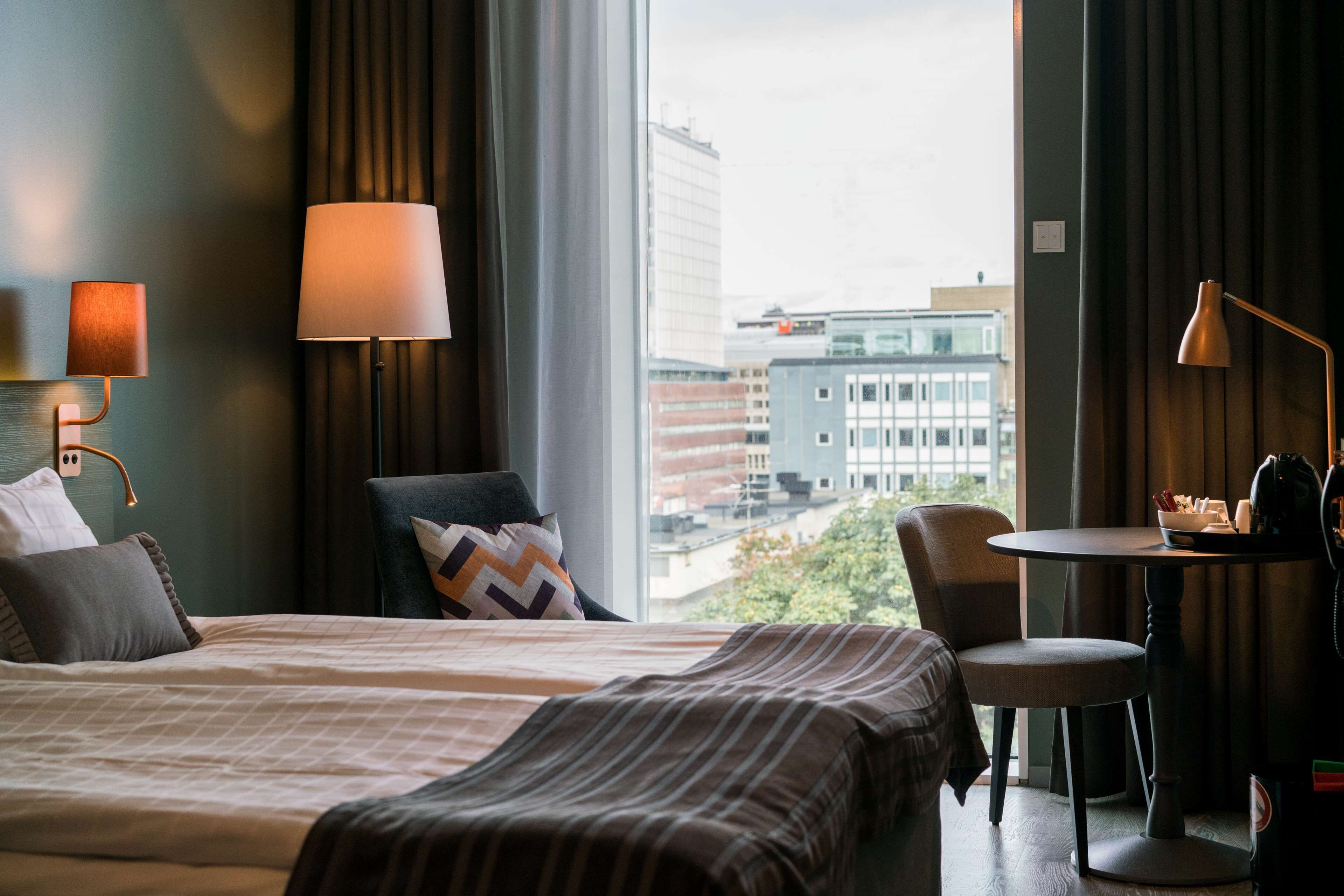 A hotel room with a bed, a chair, a small table, and a lamp. There is a window with a view of buildings outside.