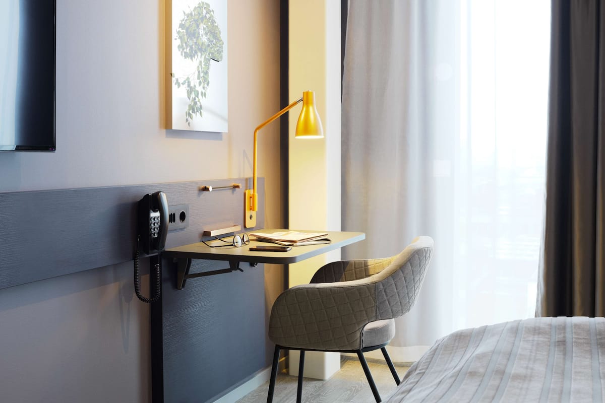 A hotel room with a bed, a desk with a telephone, a chair, a desk lamp, and a window with curtains.