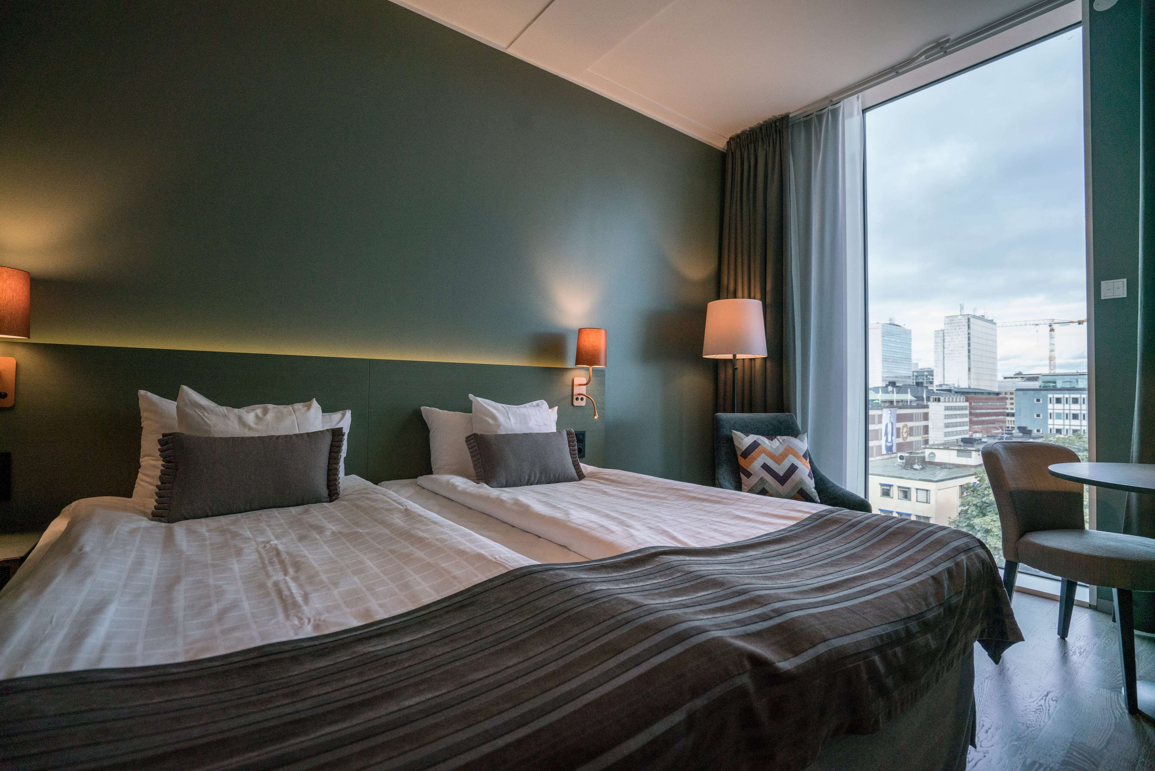 Un cuarto de hotel con una cama grande, dos lámparas de noche, vista de la ciudad a través de un ventanal amplio y una pequeña mesa junto a la ventana.