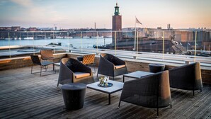 A rooftop terrace with wicker furniture, a table with drinks, and a view of a cityscape including a tower and a river. - Scandic Continental (Stockholm)