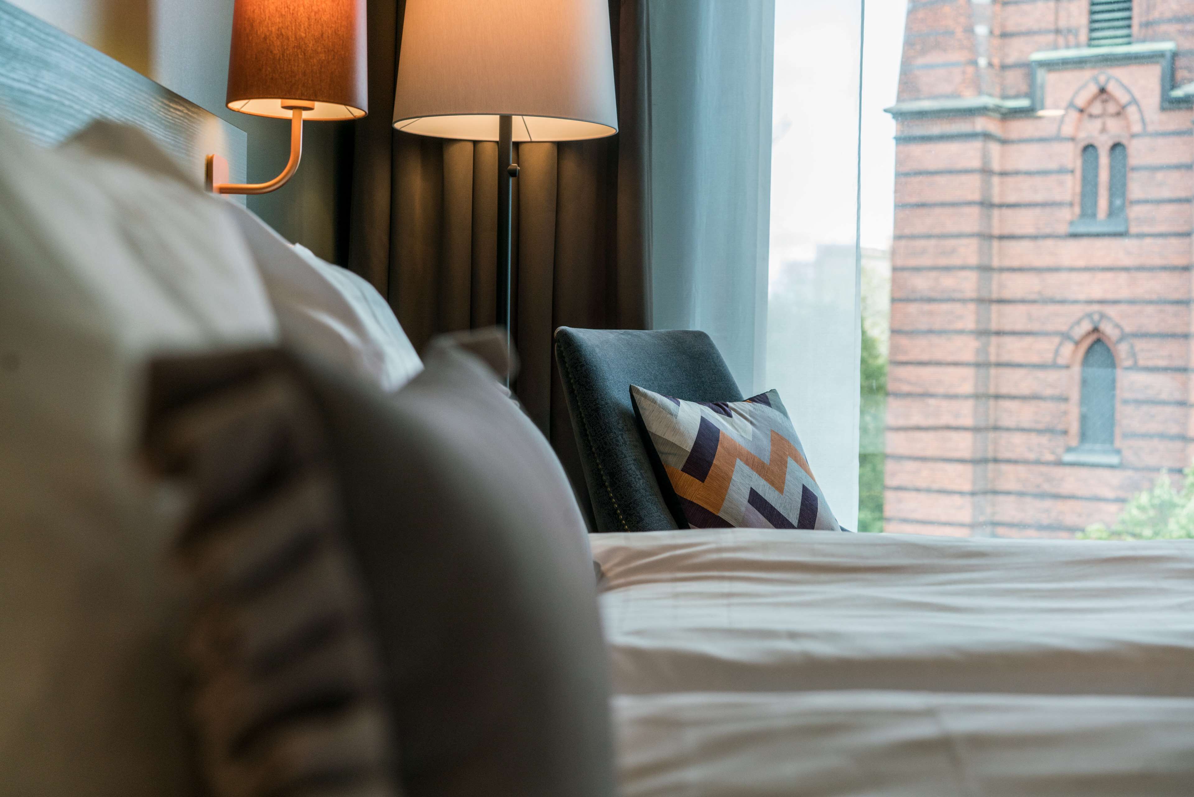 A hotel room with a bed, a lamp, a chair, and a view of a brick building.