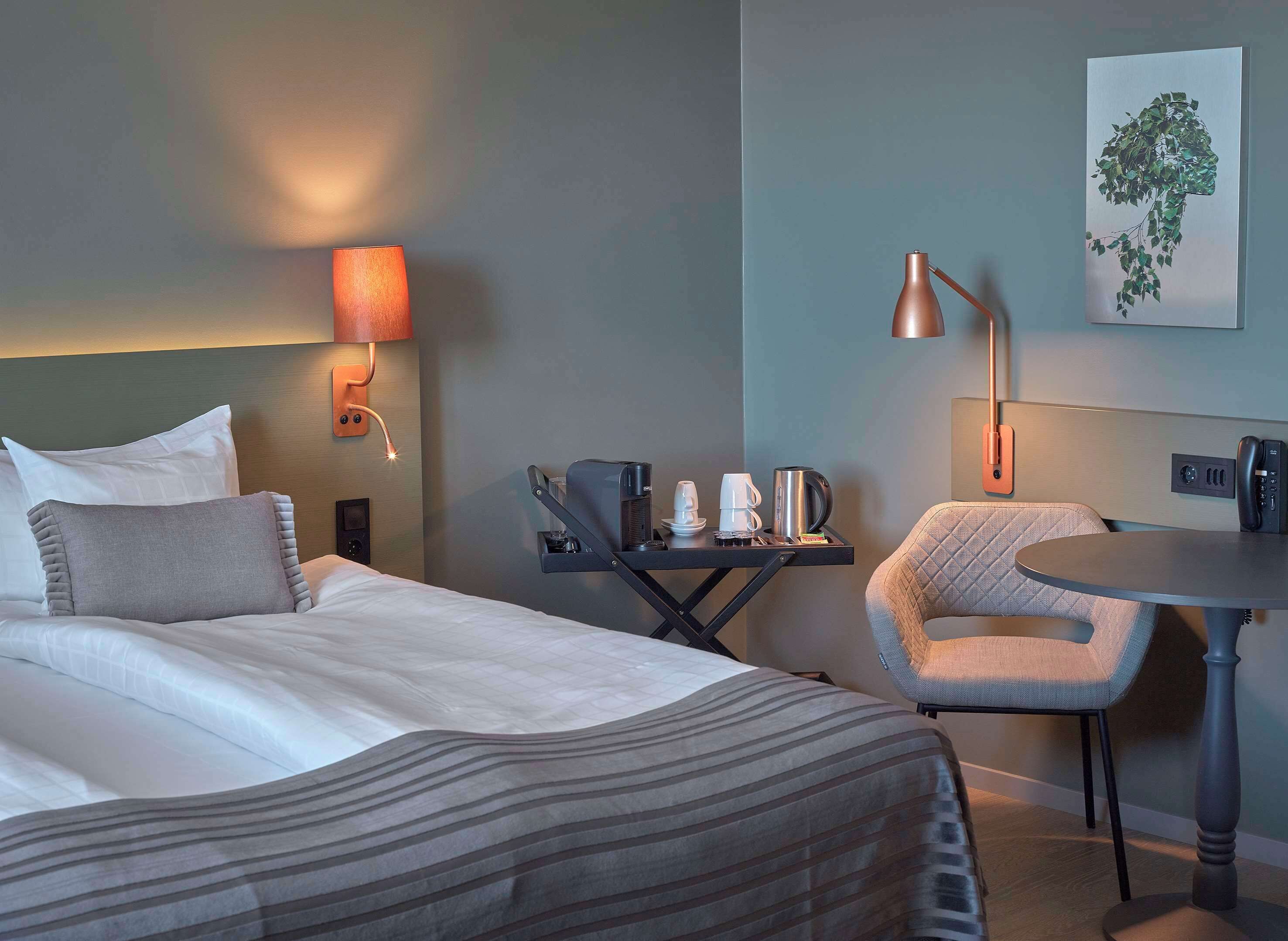 Habitación de hotel con cama, mesita de noche, silla y lámpara de pared.
