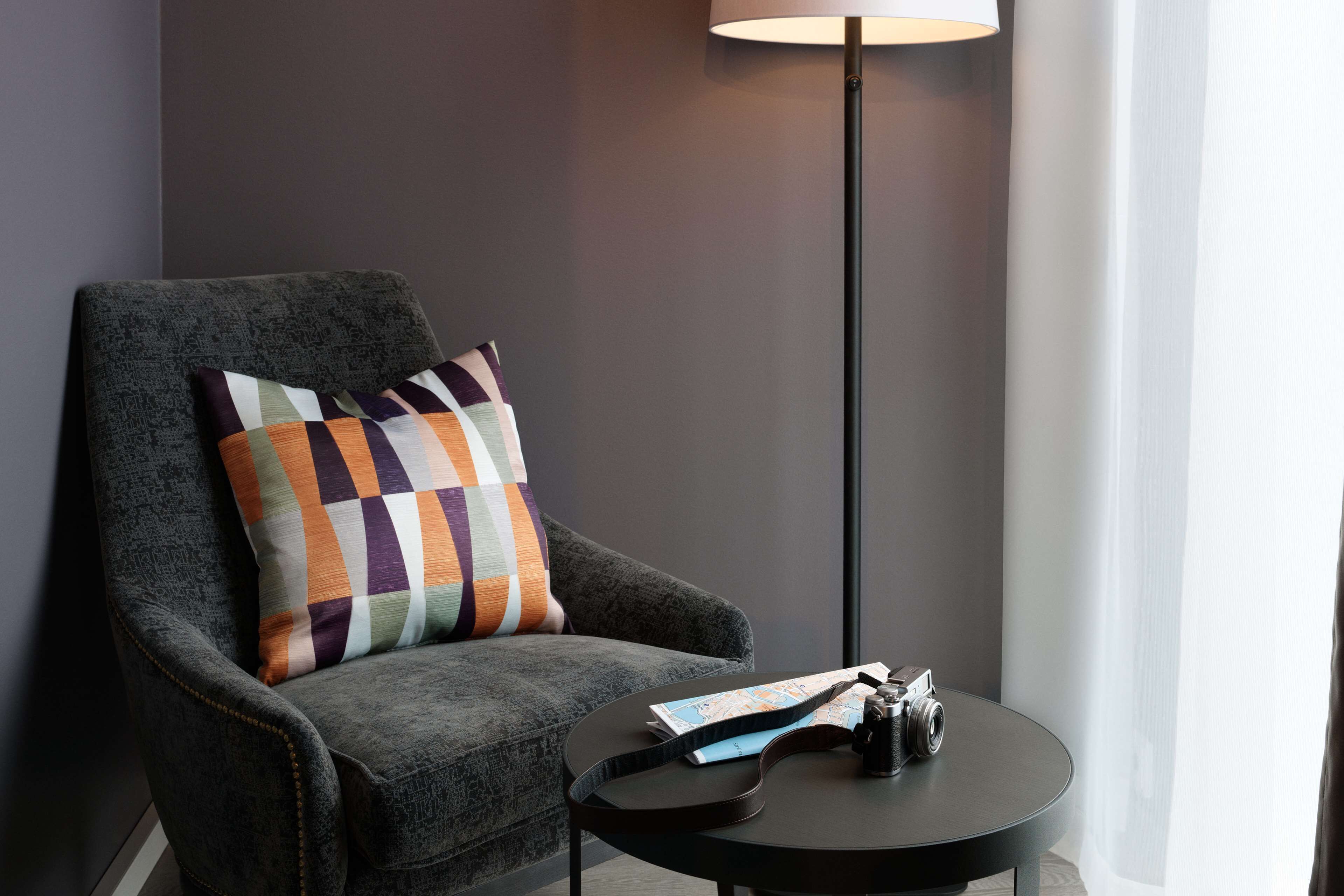 A grey armchair with a multicolored pillow, a small round table with a camera and papers, and a tall floor lamp.
