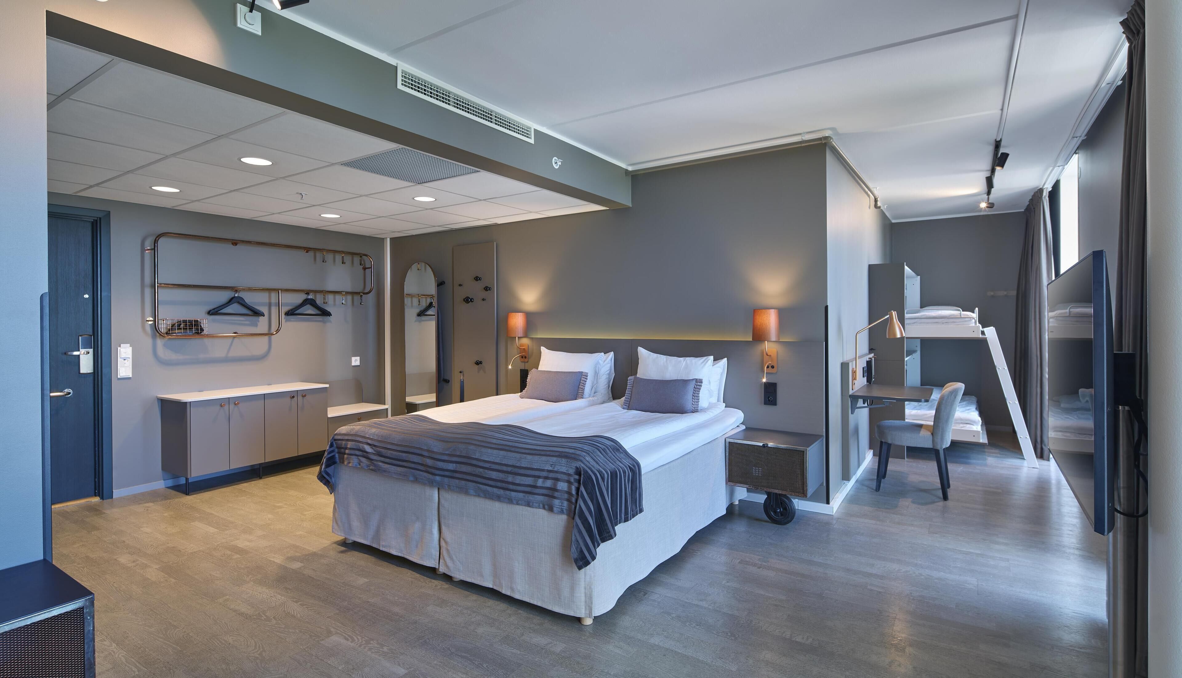 A modern hotel room with a large bed, bedside tables, a desk, and a bunk bed.
