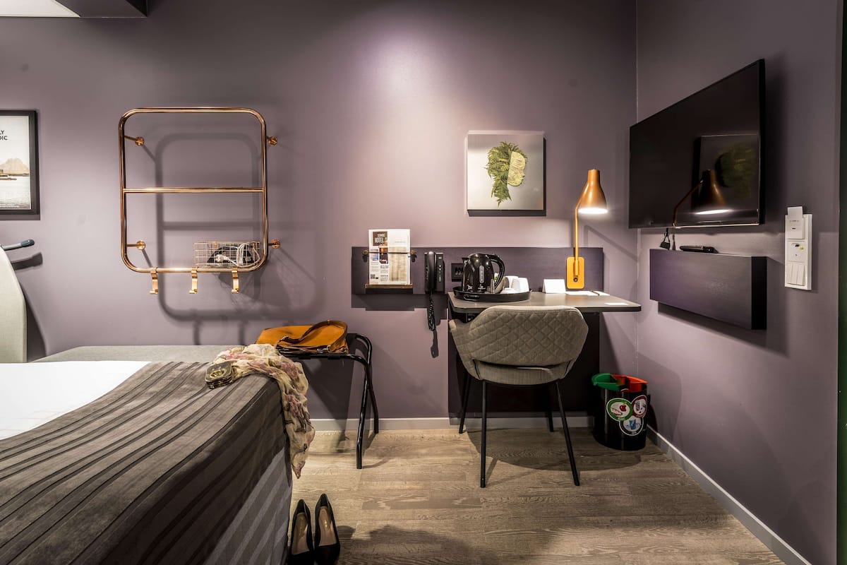 A room with a bed, desk, chair, TV, and a wall-mounted shelf with items.