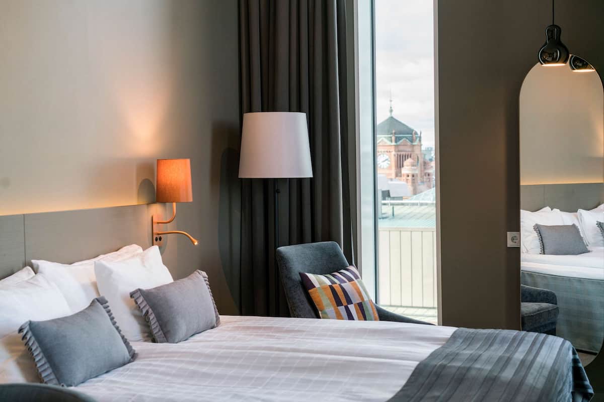 A hotel room with a large bed, a chair, a lamp, a mirror, and a view of a building.