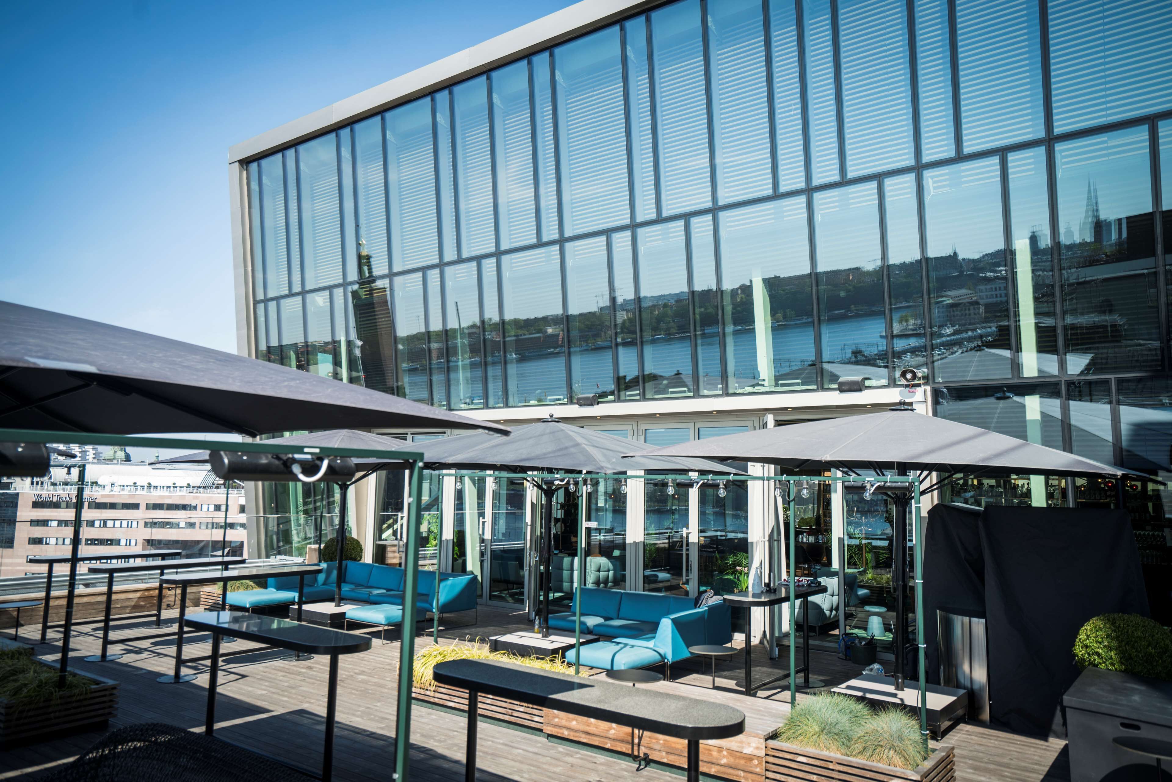En takterrass med utomhusmöbler, utsikt över en modern byggnad och en vattenyta i bakgrunden.