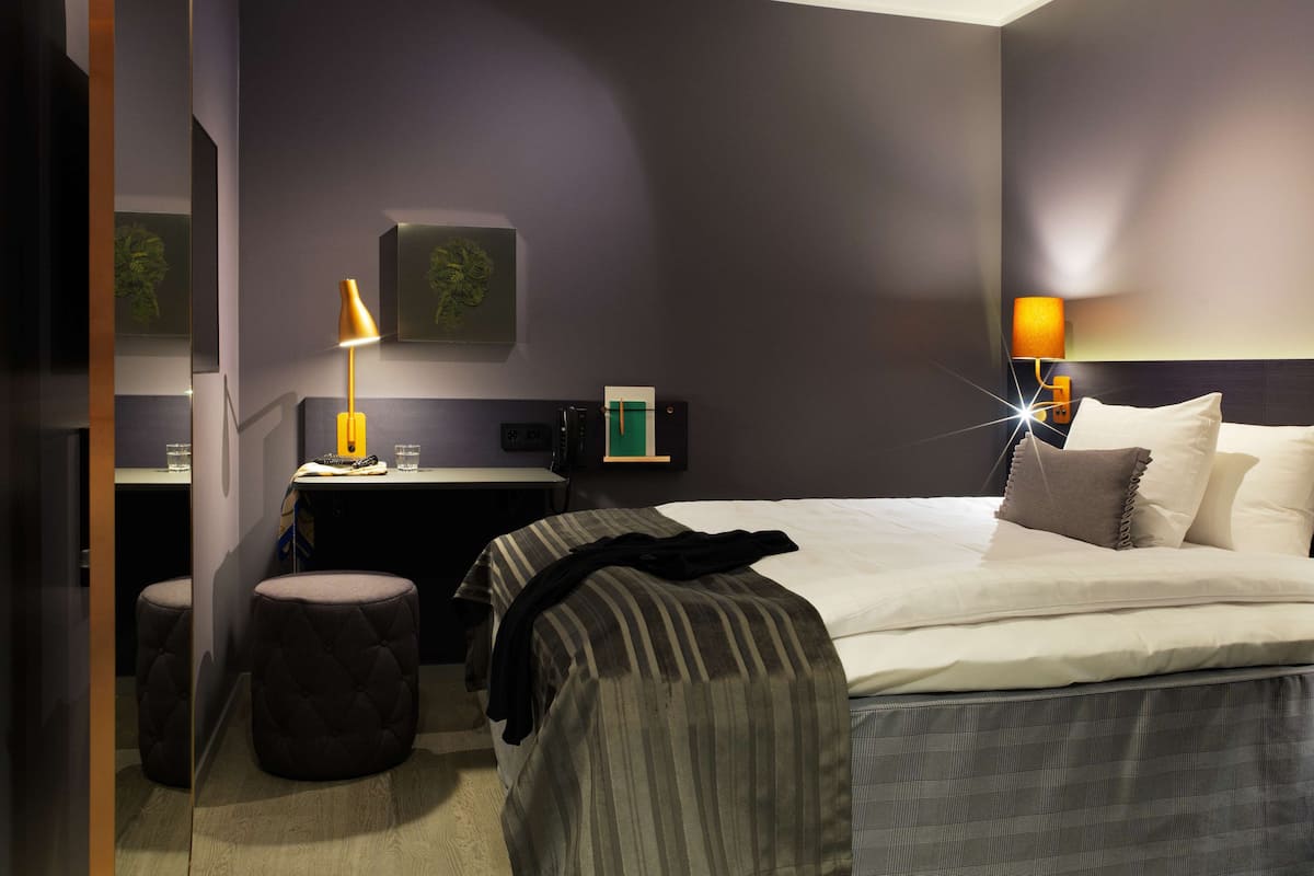 A modern hotel room with a bed, two ottomans, a desk, and two wall-mounted lamps.
