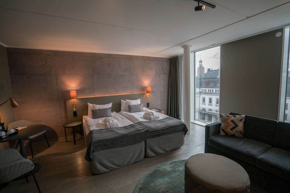 A modern hotel room with a large bed, a sofa, a small table, and a view of a cityscape.