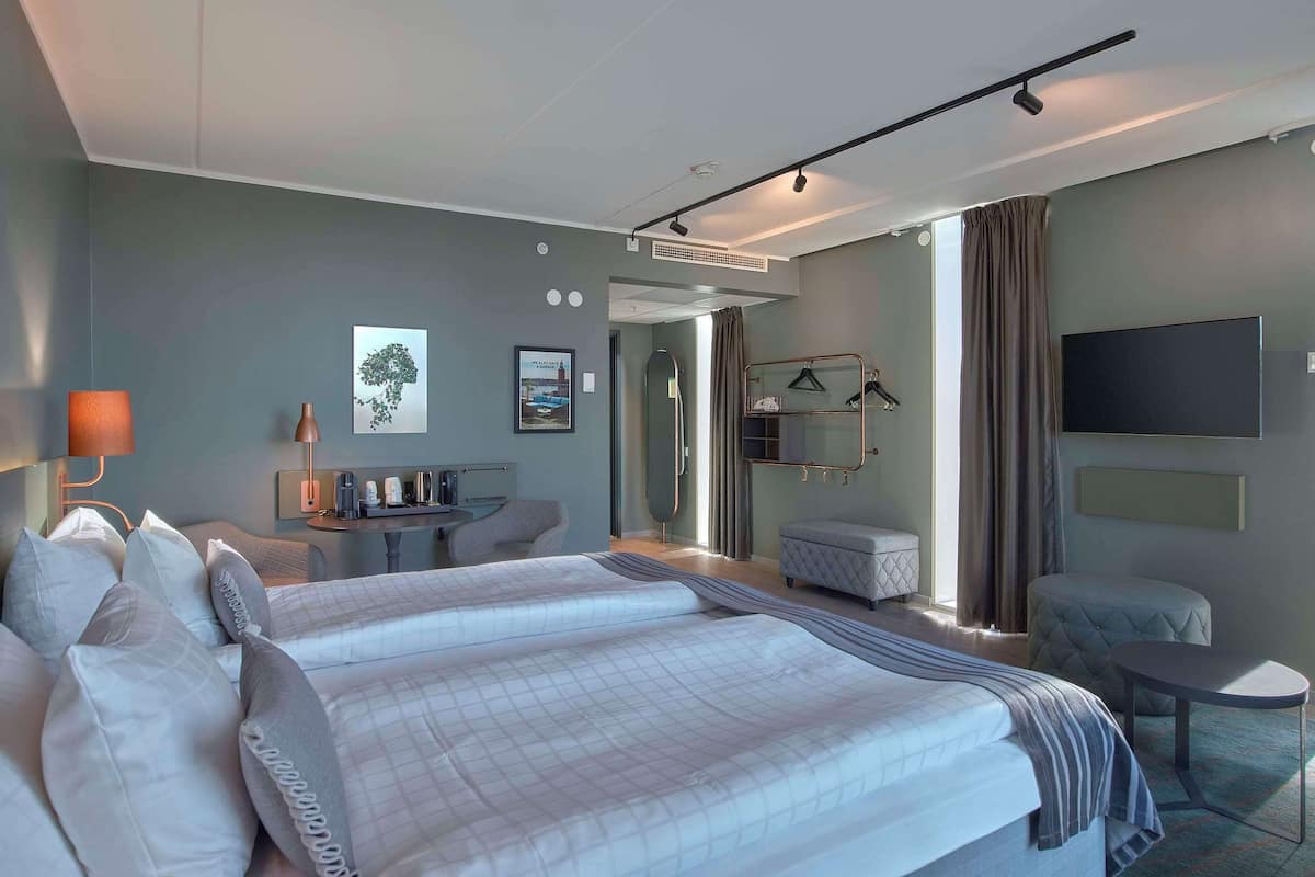 A hotel room with a bed, bedside tables, a desk, a chair, a TV, and a window with curtains.