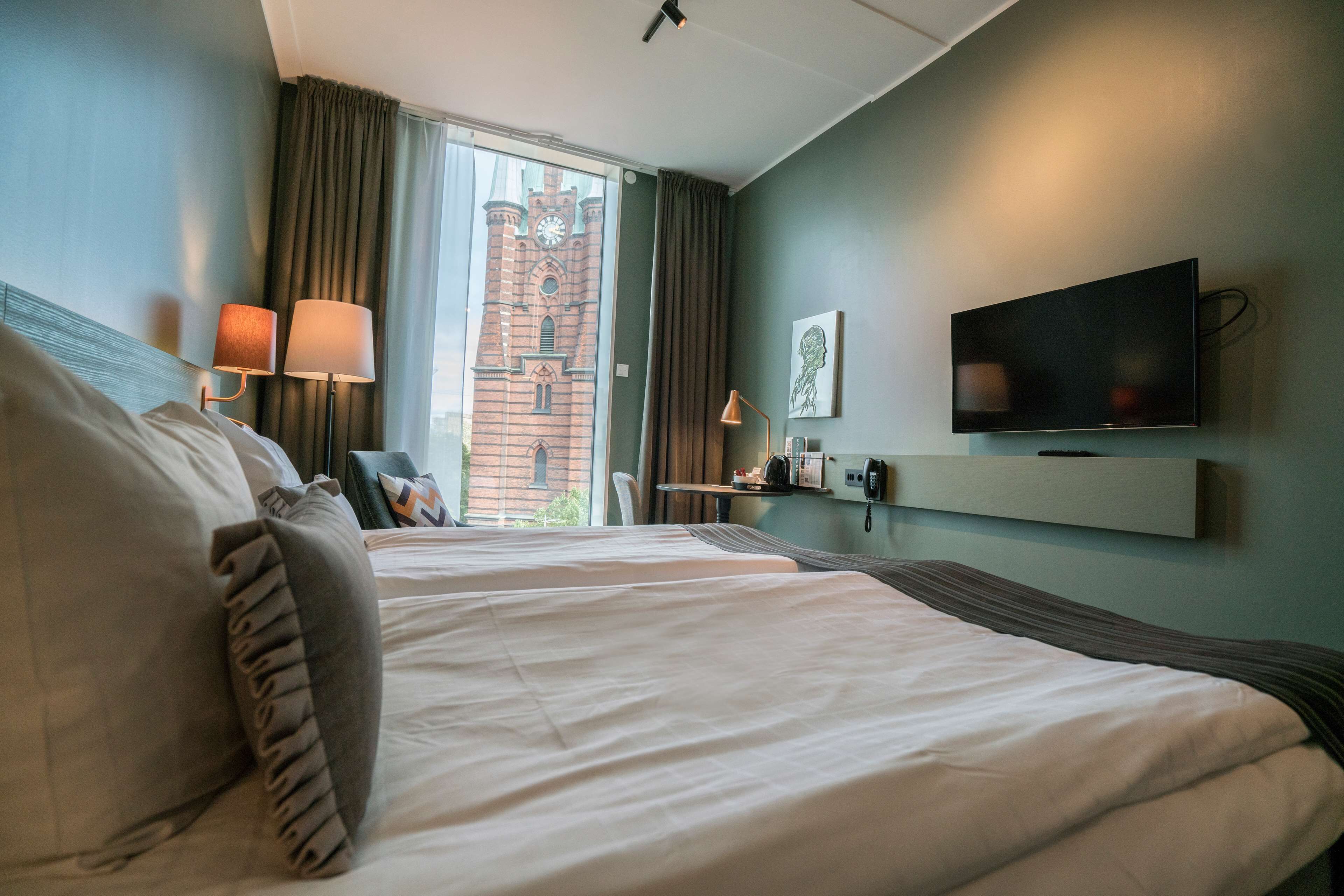 A hotel room with two beds, a TV, a window with curtains, and a brick building view.