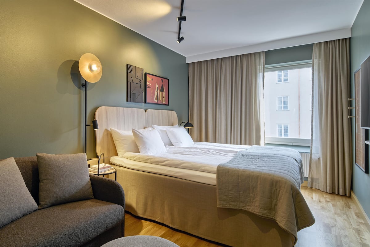 Ein Hotelzimmer mit Bett, Sofa, Nachttisch, Lampe und einem Fenster mit Vorhängen.
