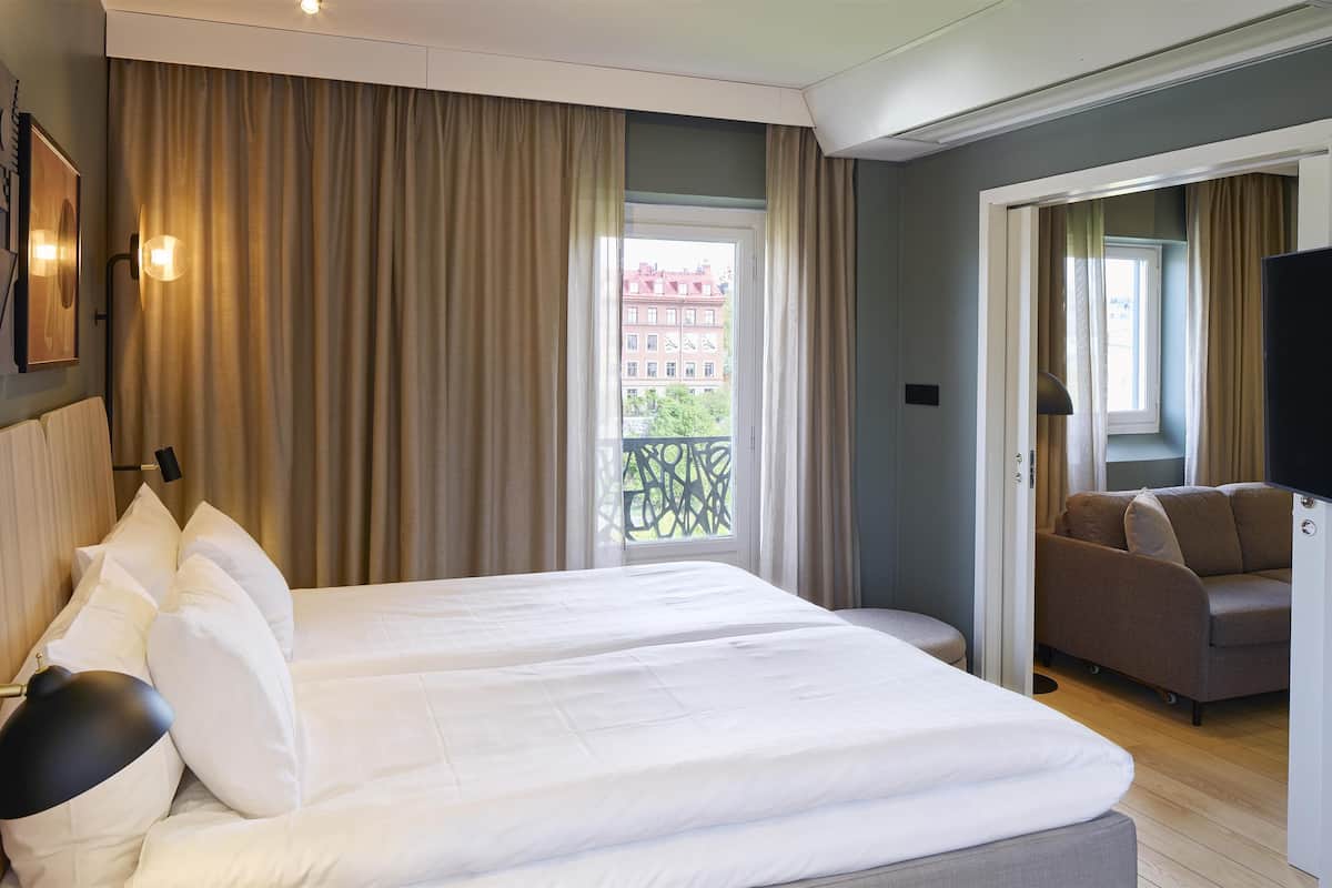 Ein Hotelzimmer mit einem großen Bett, einem Fernseher, einer Couch und einem Balkon mit Blick auf Gebäude.