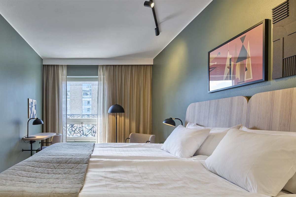 Ein Hotelzimmer mit einem großen Bett, einem Fernseher, einem Schreibtisch und einem Fenster mit Vorhängen.