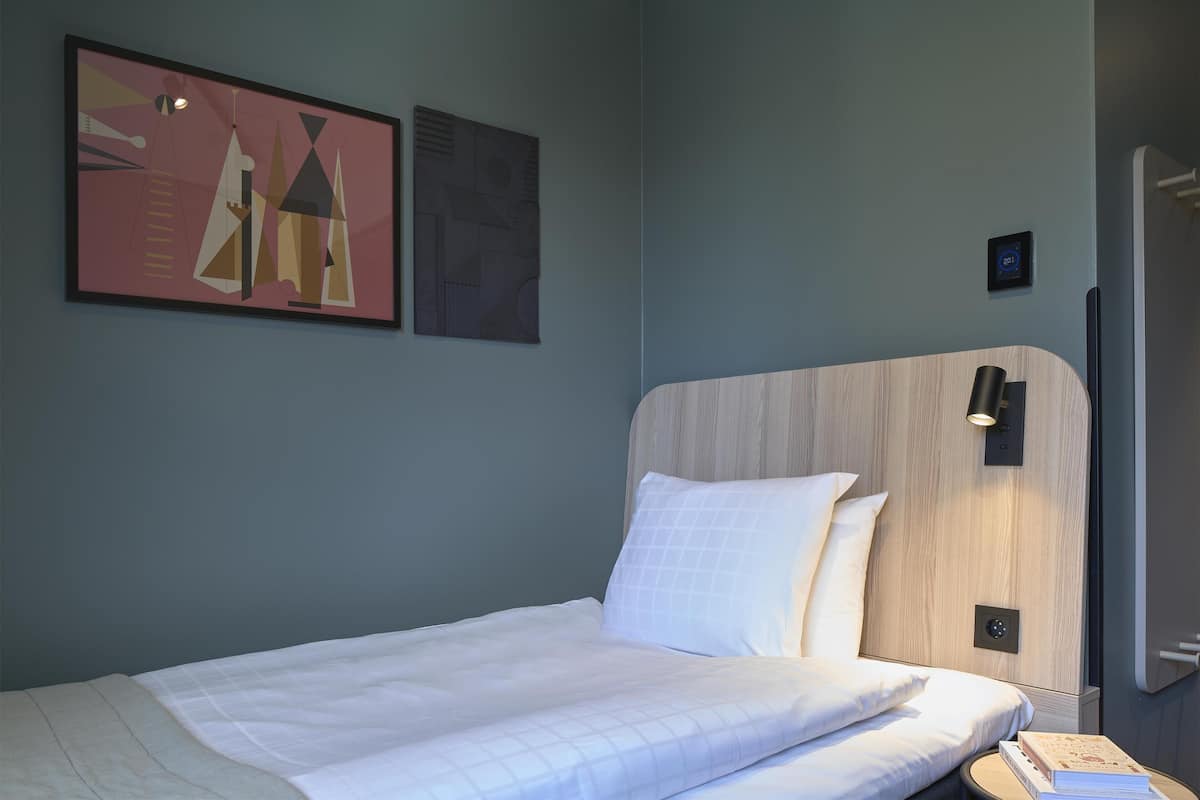 Ein ordentlich bezogenes Bett mit weißen Leinen, ein Kopfteil mit heller Holzoptik und eine wandmontierte Lampe.