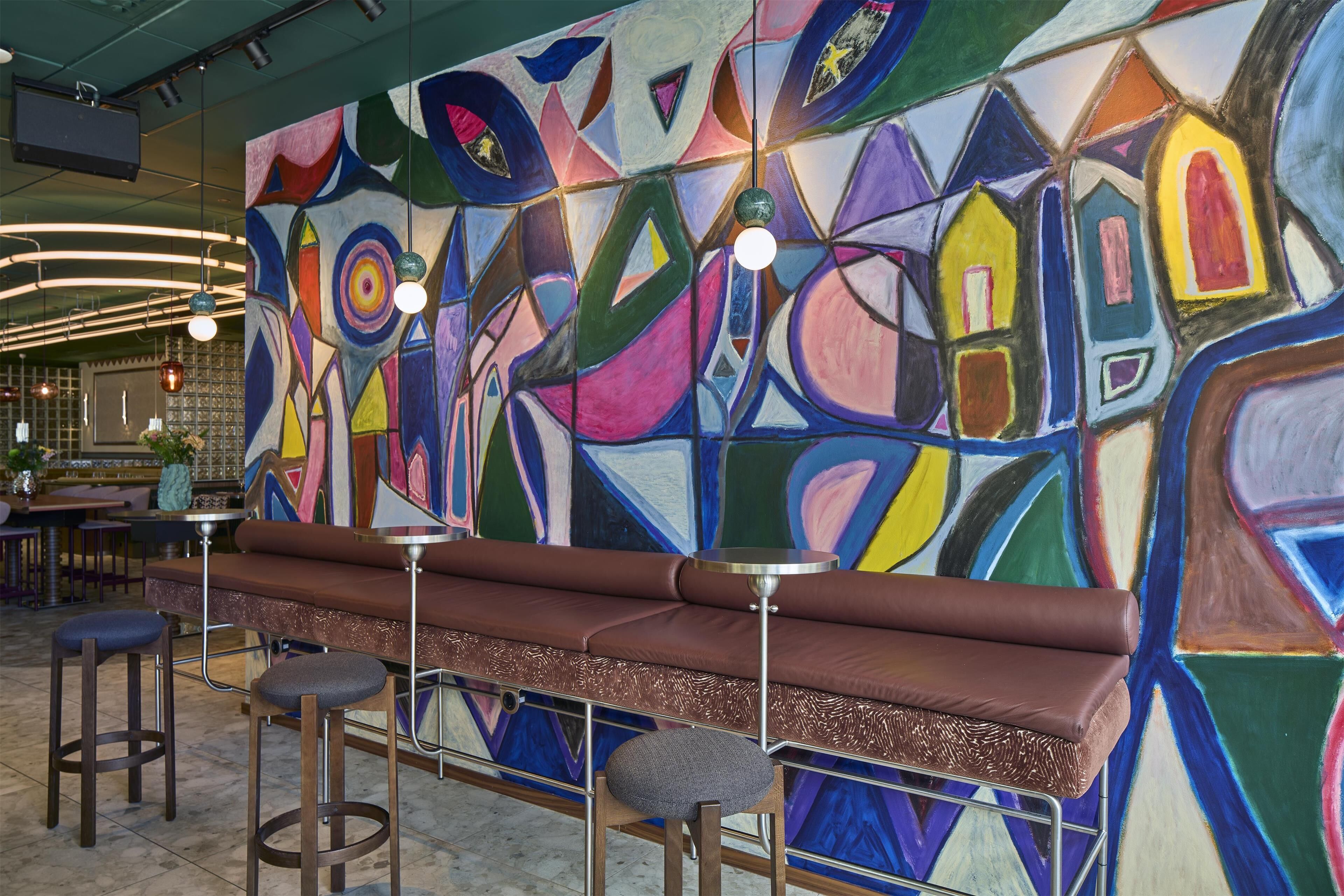 Een bar met een kleurrijk abstract schilderij, een bruine met kussens beklede bank en twee barkrukken.