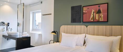 Una habitación de hotel moderna con una cama grande, un baño con lavamanos y espejo, y obras de arte en las paredes.