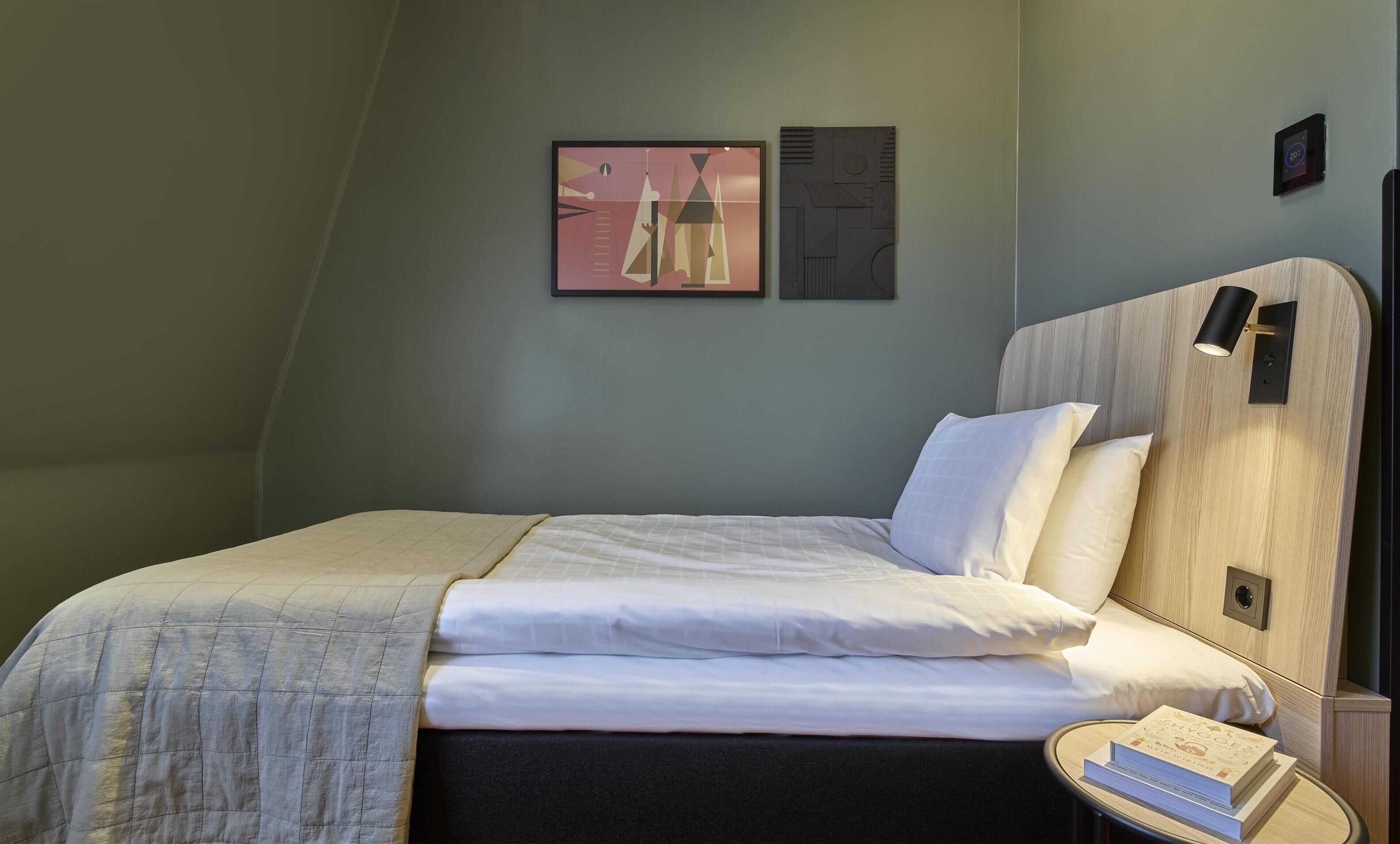 A neatly made bed with white linens and a beige blanket, a bedside table with a book, and a wall-mounted light fixture.