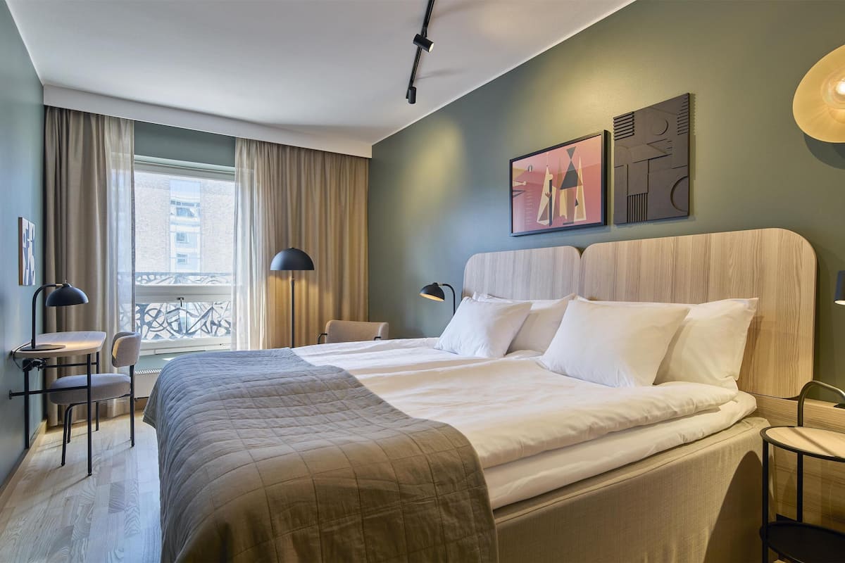 Ein Hotelzimmer mit einem großen Bett, einem Schreibtisch mit Stuhl, einer Stehlampe und einem Fenster mit Vorhängen.