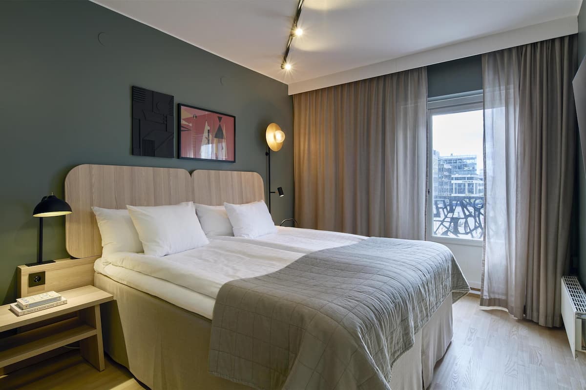 Ein modernes Hotelzimmer mit einem großen Bett, einem Nachttisch, einer Lampe und Blick auf die Stadt durch das Fenster.