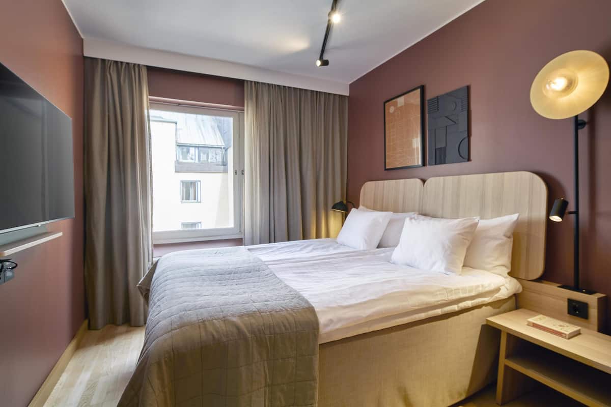 Ein Hotelzimmer mit einem großen Bett, einem Flachbildfernseher, einem Nachttisch mit Lampe und einem fenster mit Vorhängen.