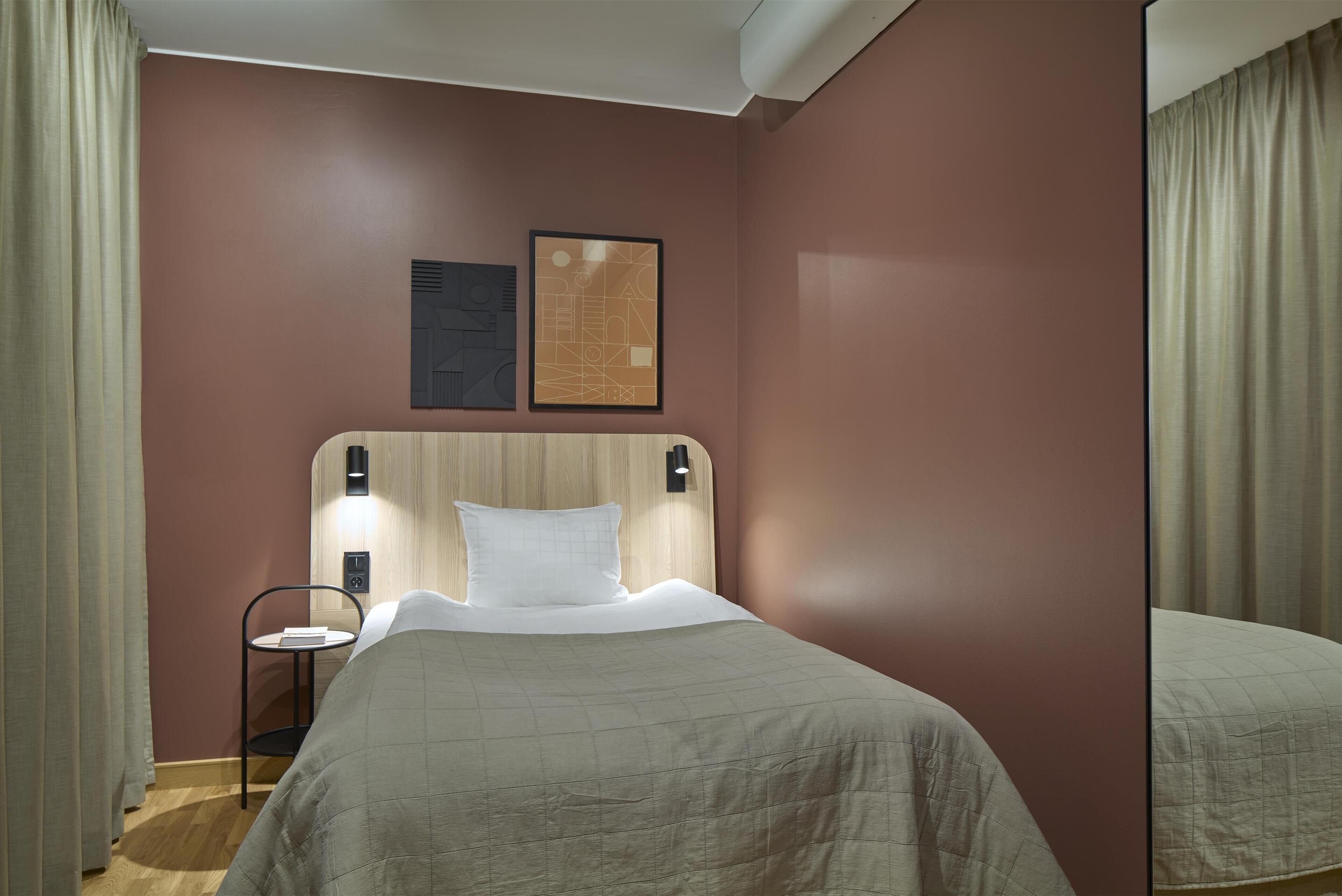 A hotel room with a bed, a nightstand, a wall-mounted lamp, and two framed pictures.