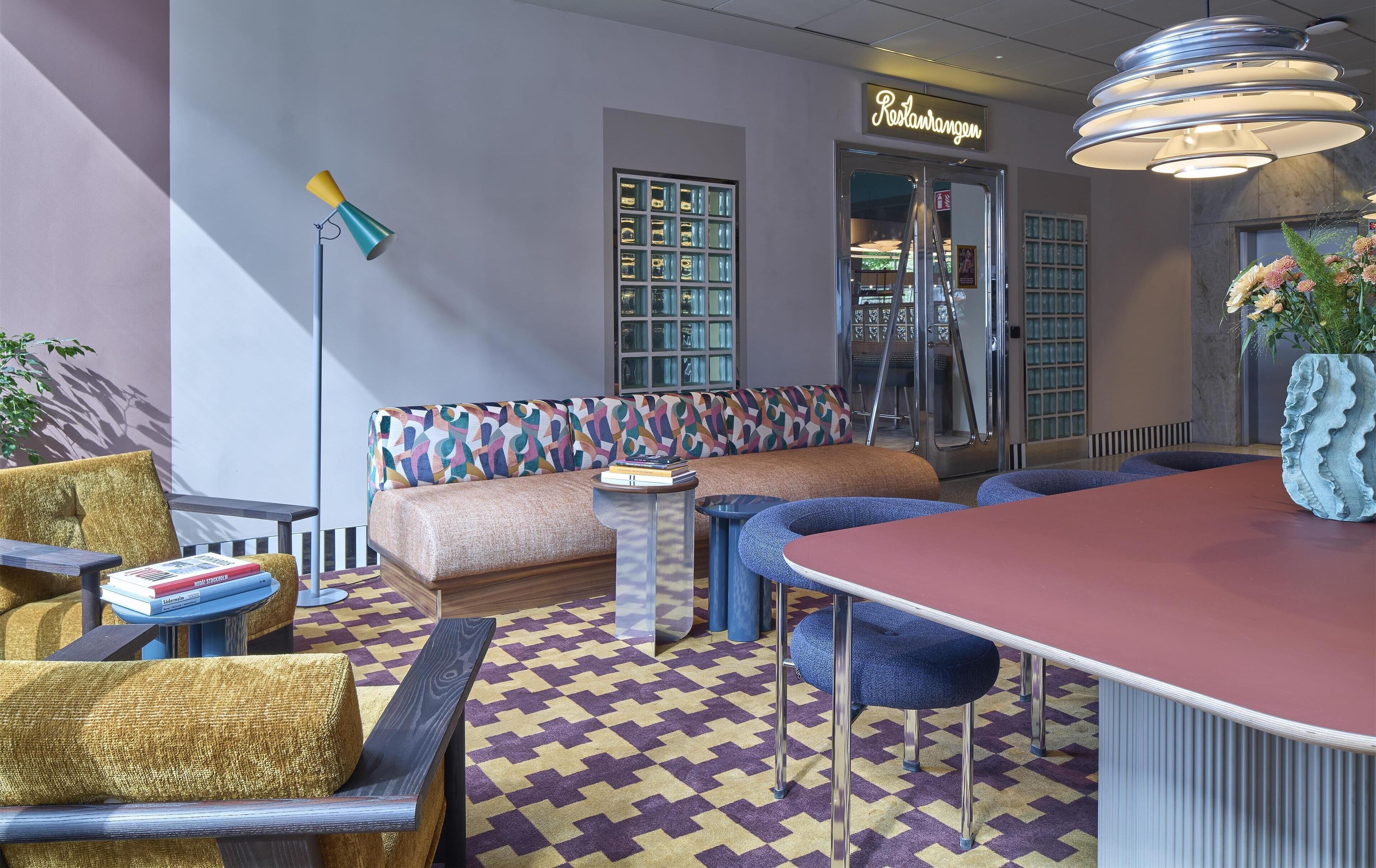 A modern lounge area with a patterned carpet, a seating area with a colorful sofa, a bar with stools, and a decorative plant.