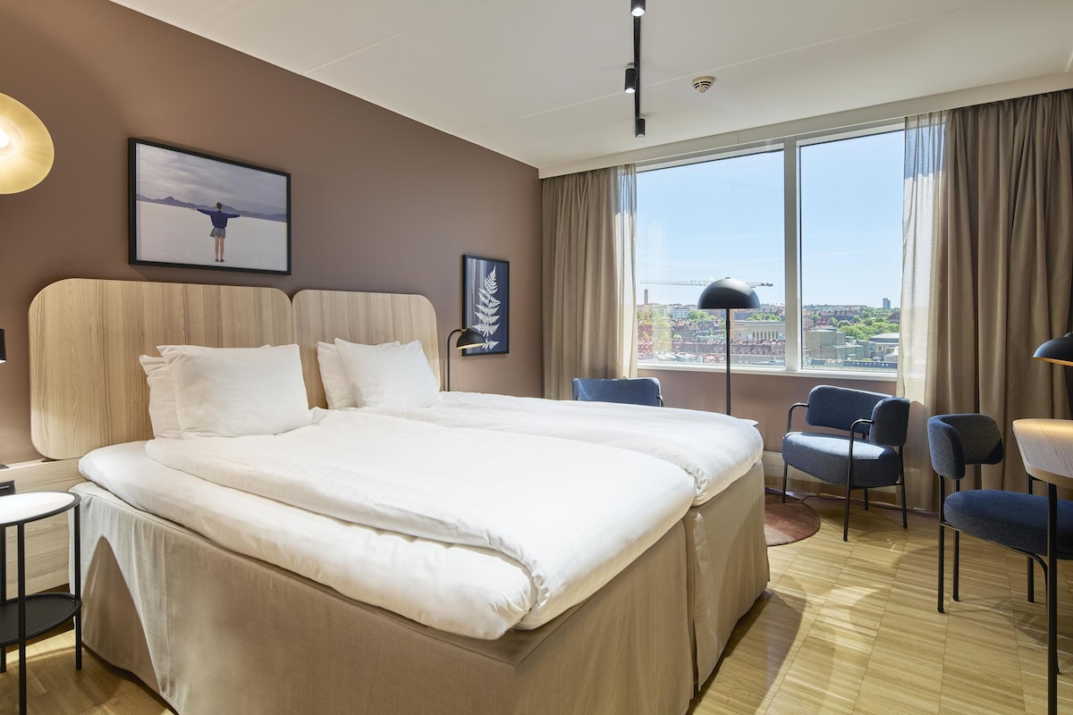 Habitación de hotel con una cama grande, dos sillas, un escritorio y ventana con vistas a la ciudad.