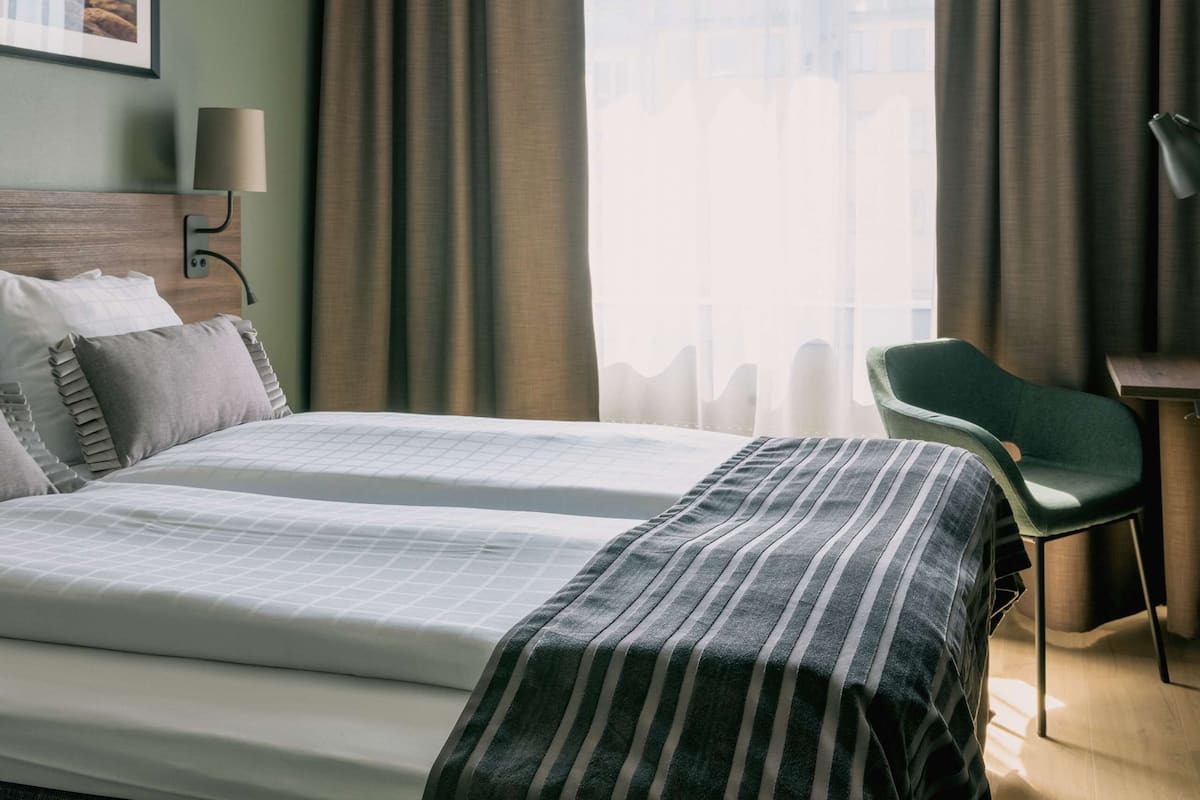 A hotel room with two beds, a desk, a chair, a lamp, and a large window with curtains.