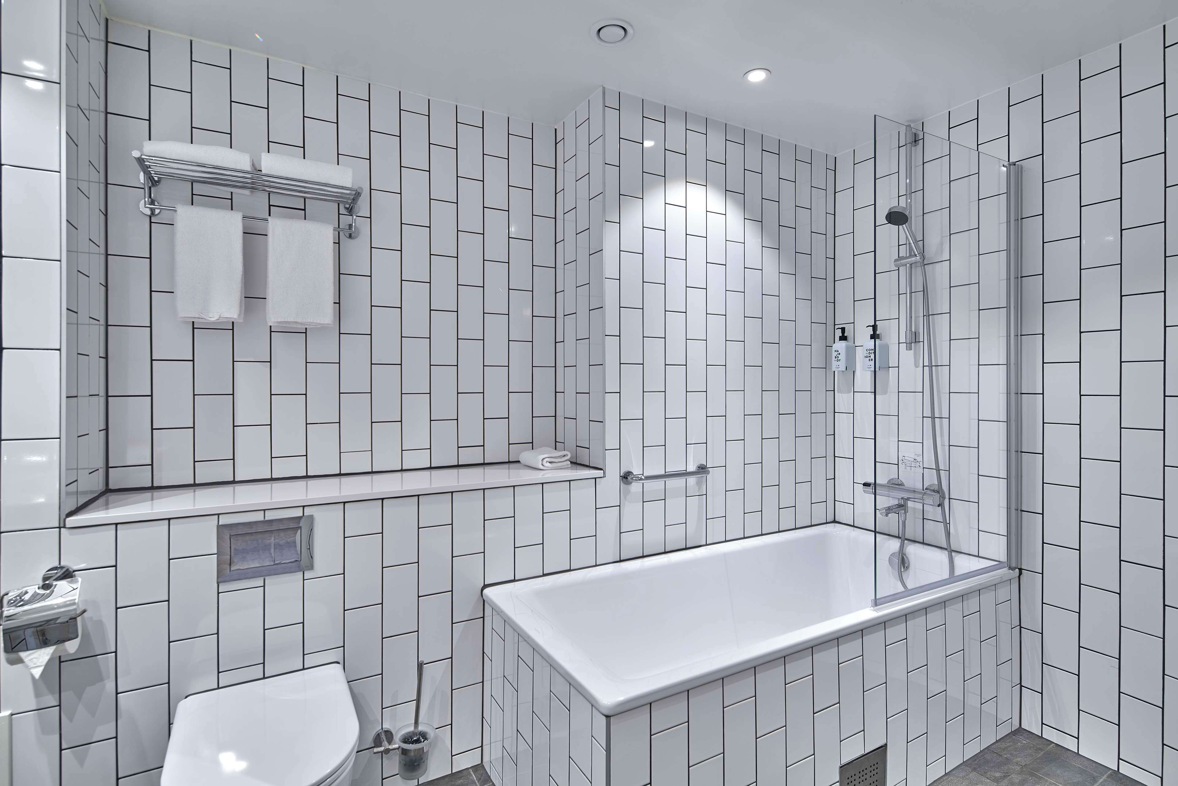 Un baño moderno con pared de azulejos blancos, una bañera rectangular, un inodoro y una ducha.