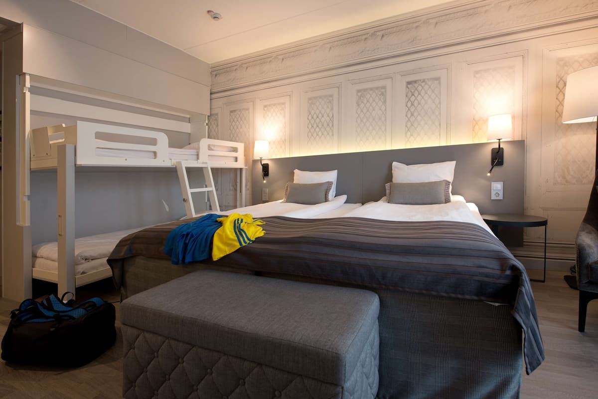 A hotel room with a bunk bed, a desk, a chair, and a bedside table with a lamp.