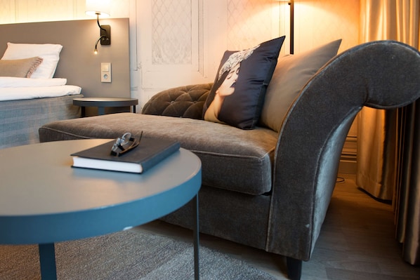 A modern hotel room with a grey sofa, a round table with a book, a bedside lamp, and a bed with white linens. - Scandic Rubinen (Gothenburg)