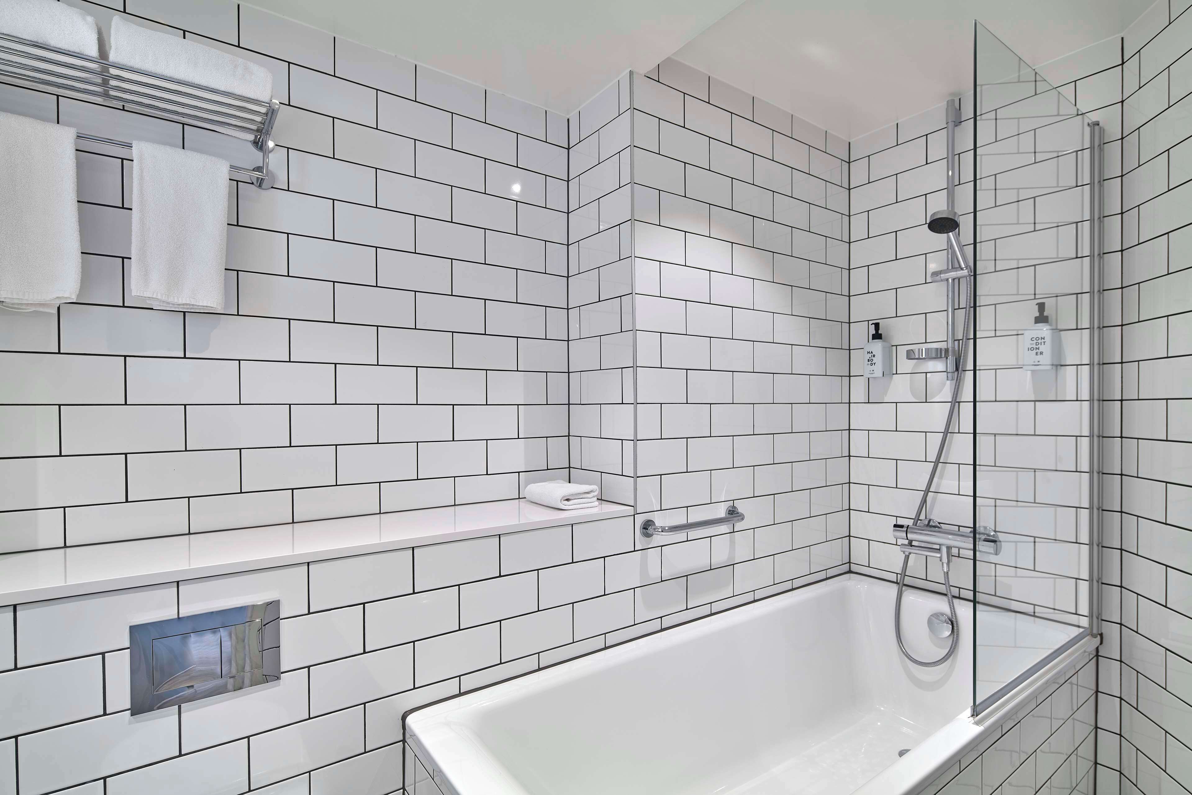 Une salle de bain moderne avec des carreaux de céramique blancs rectangulaires, une douche à parois de verre, une baignoire blanche et un porte-serviettes avec deux serviettes.