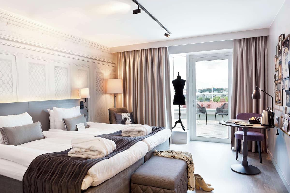A hotel room with a large bed, a desk with a chair, a mannequin, and a view of the city through the window.