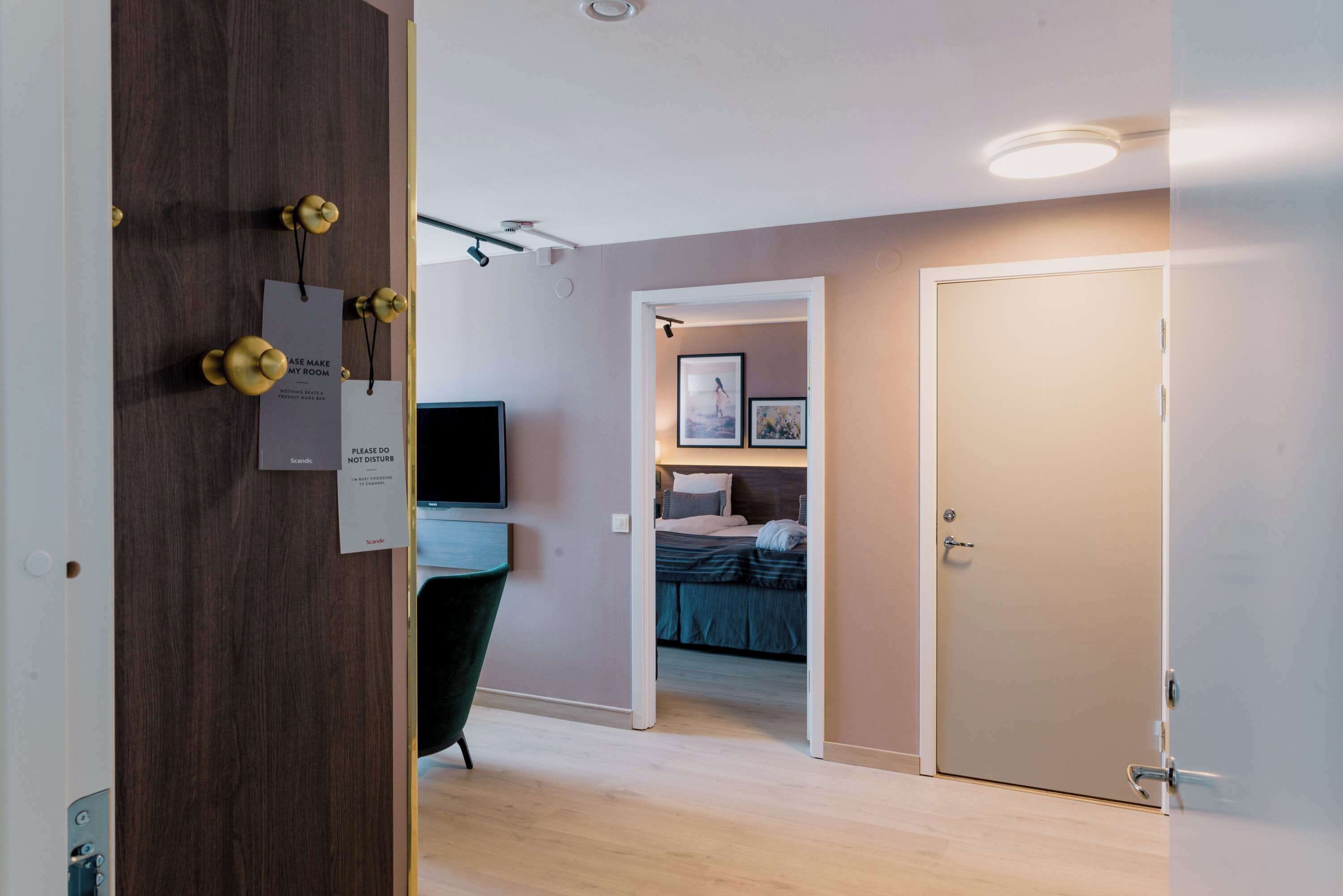 Habitación de hotel con cama, escritorio y televisor. Hay letreros en la puerta y un cuadro en la pared.