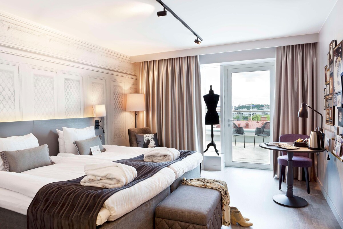 Habitación de hotel con una cama grande, un escritorio con una silla, un maniquí y vistas de la ciudad a través de la ventana.