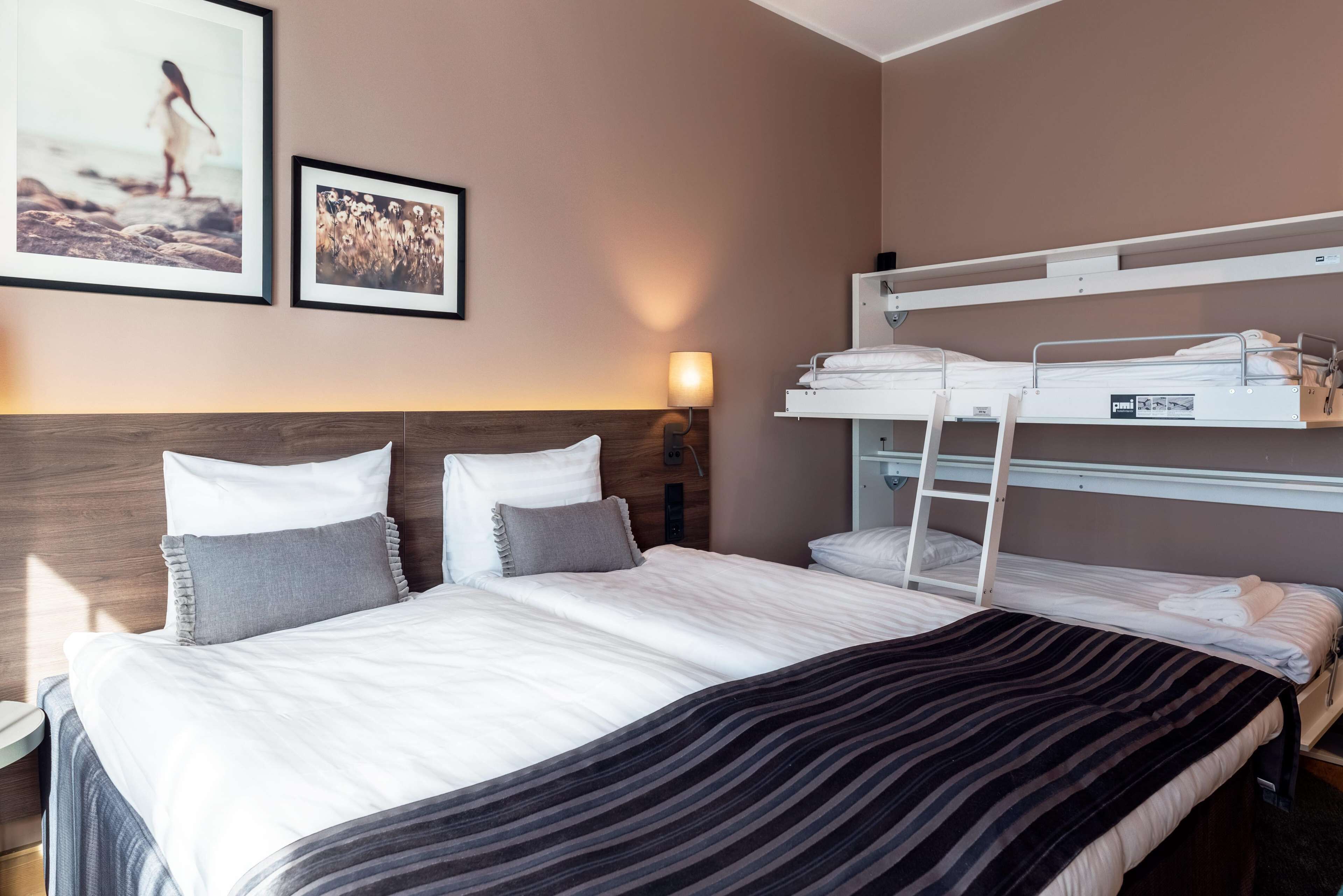 Habitación de hotel con literas, una cama grande con almohadas, una mesita de noche y cuadros enmarcados en la pared.