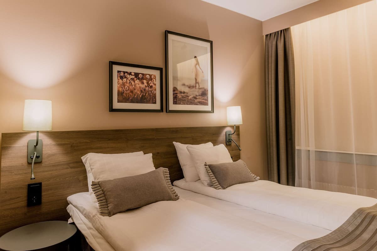 A hotel room with a wooden headboard, two beds with white and brown bedding, two wall-mounted lamps, a small round table, and two framed pictures on the wall.