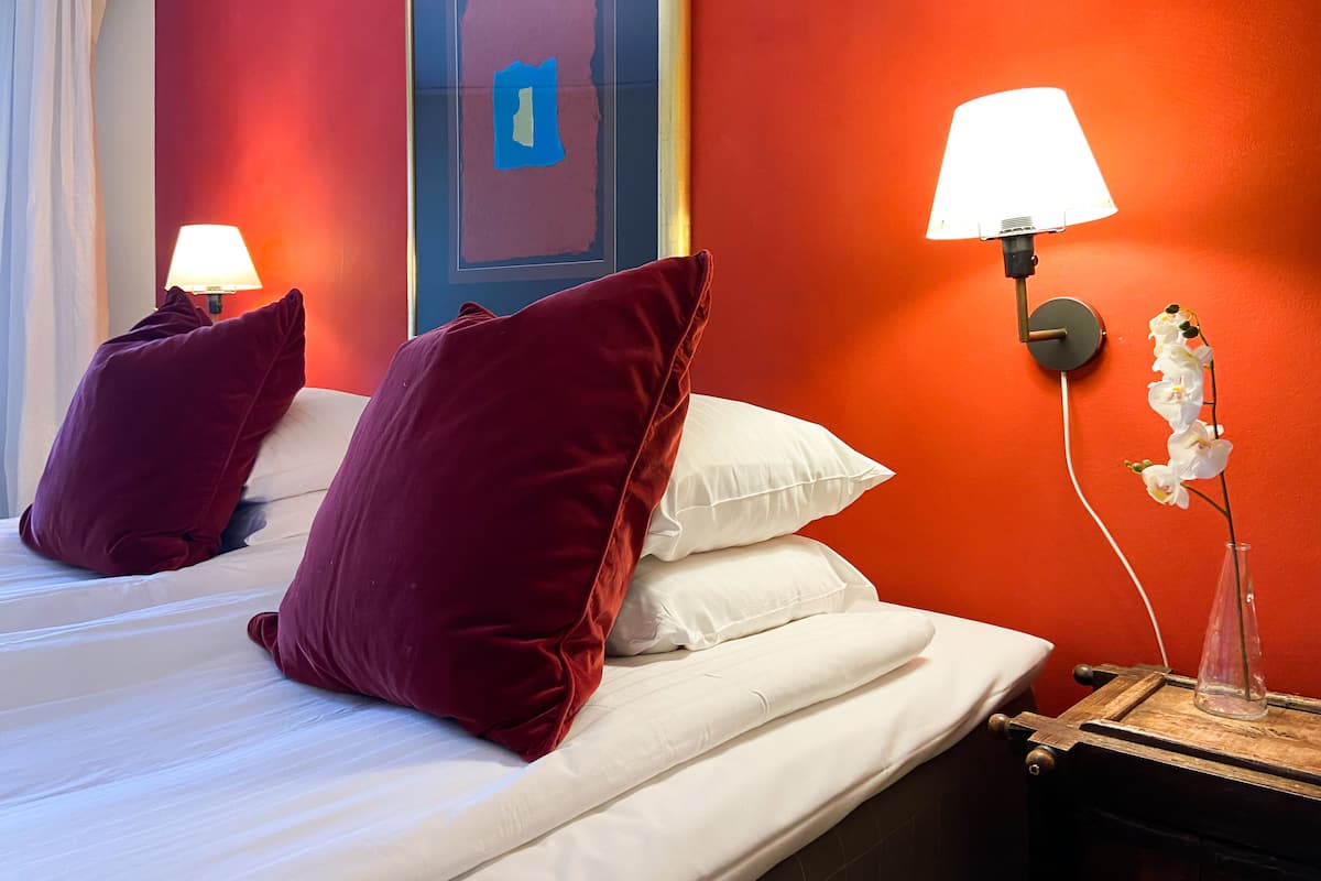 A hotel room with a red wall, a bed with white and maroon pillows, a nightstand with a lamp, and an orchid plant.